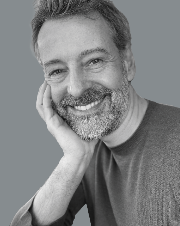 Smiling man with a beard and gray hair, resting his cheek on his hand. Wearing a ribbed sweater against a gray background. 