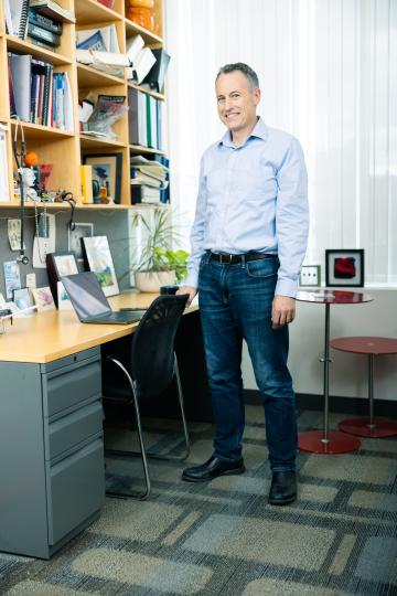 Man smiling in an office, wearing a light blue shirt and jeans. He stands by a desk with a laptop, bookshelves filled with items, and framed pictures.