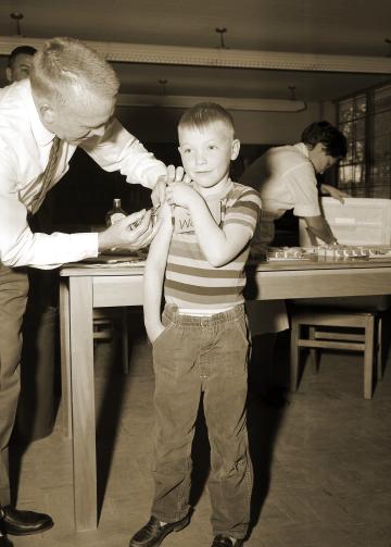 In a historic, sepia-toned image, a young boy in striped shirt receives a vaccination from an older man in a tie.