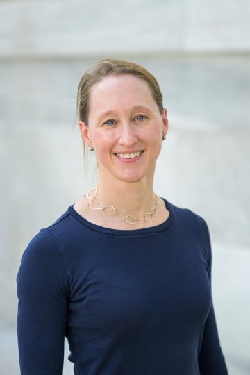 A woman is smiling in a navy blue top with a chain necklace, standing against a light blurred background.