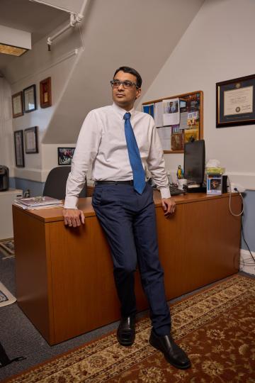 A man in a white shirt and blue tie leans casually against a wooden desk in an office. The room has framed certificates and a cluttered noticeboard.