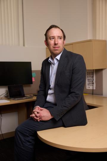 A man wearing a suit sits on the edge of a desk in a modern office, looking thoughtfully to the side. A laptop and monitor are visible in the background.