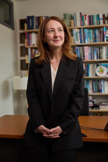 A woman in a black suit stands confidently in an office, surrounded by bookshelves. 