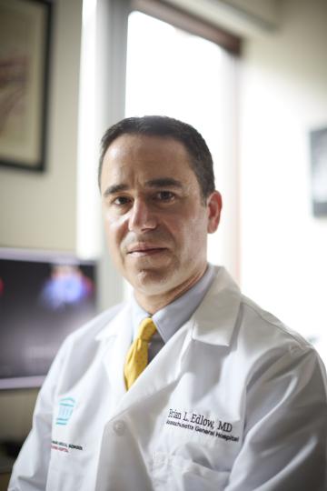A portrait of a man in a white coat with a serious expression. A computer monitor with an MRI scan is blurred in the background.