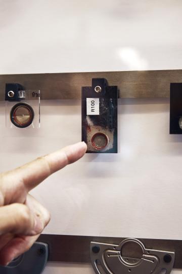 A finger points to a small, rectangular metal object labeled "R100" mounted on a display board with other metal components.
